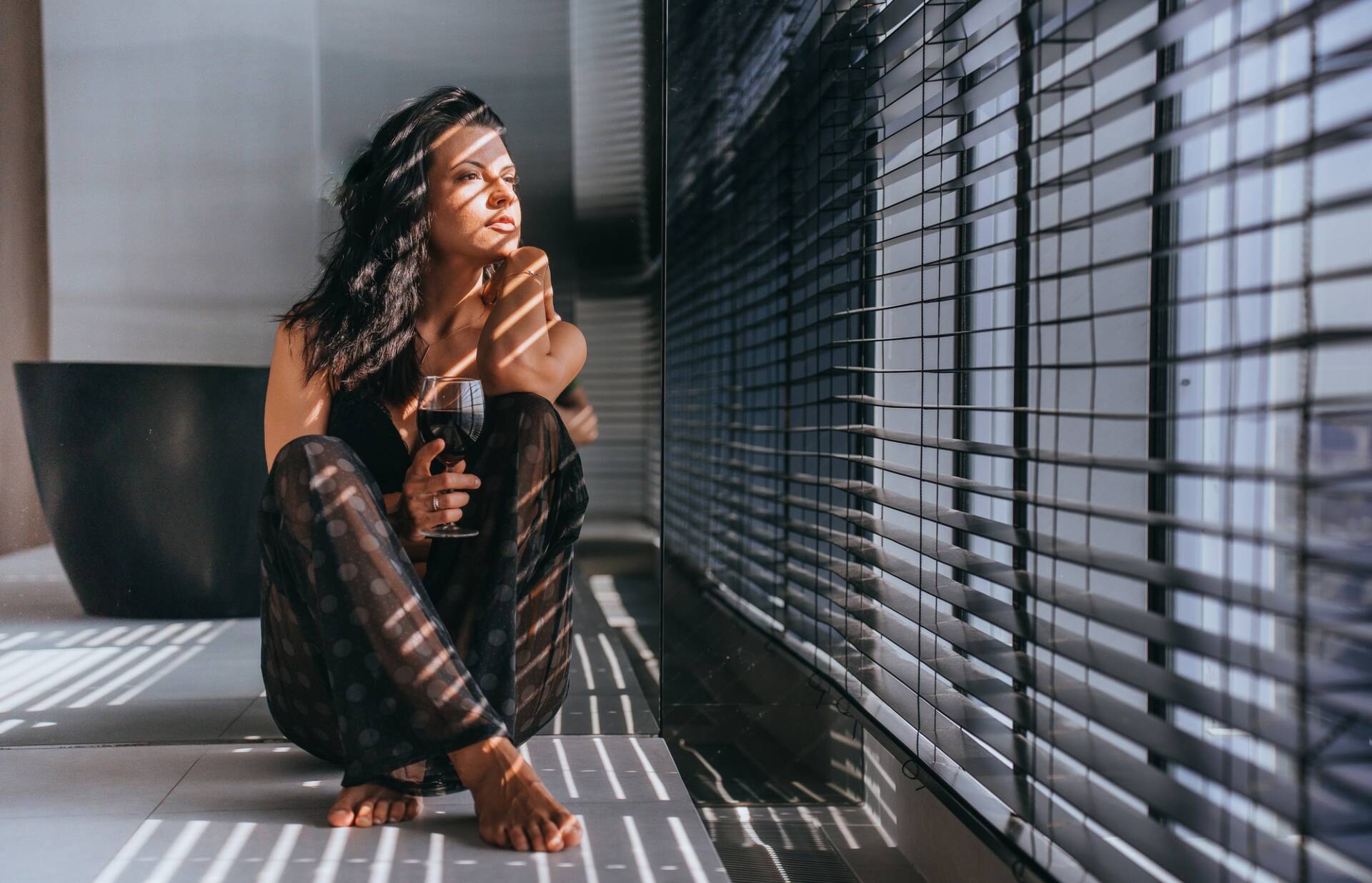 Mujer contemplativa con copa de vino sentada junto a la ventana, bañada por la luz del sol filtrada a través de las persianas - Puertas y ventanas de pvc Termprotect