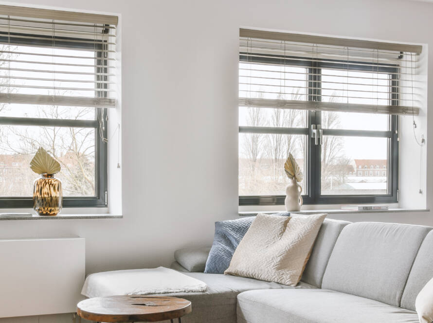 Ventana de PVC moderna con persianas, integrándose en una sala minimalista con sofá gris y mesas auxiliares.