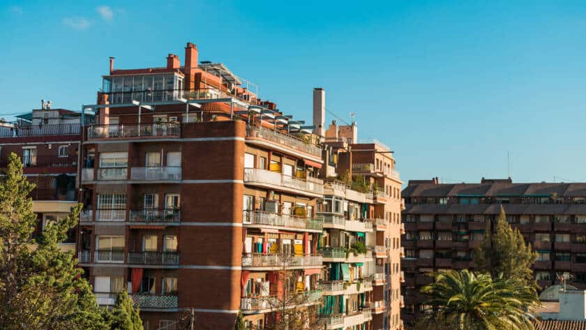Fachada de un edificio residencial de ladrillo visto en Barcelona, con varios balcones y terrazas. El cielo es azul y despejado. La imagen representa una vivienda típica con potencial para mejoras de aislamiento térmico.